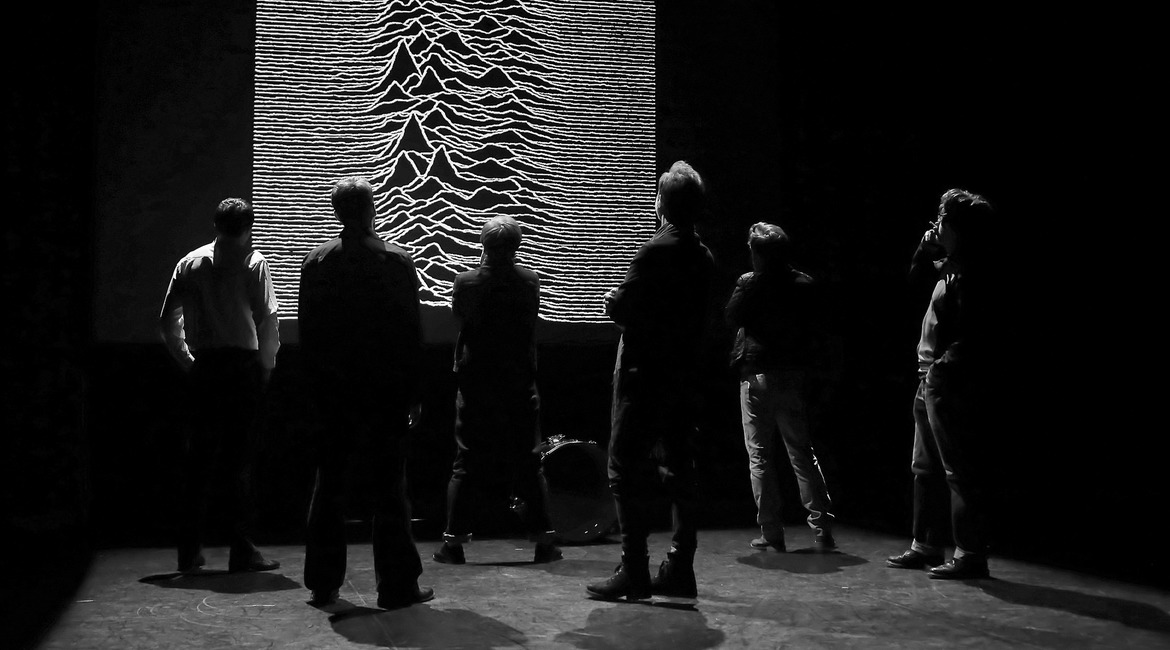 A production shot of actors onstage in front of Joy Division artwork in New Dawn Fades
