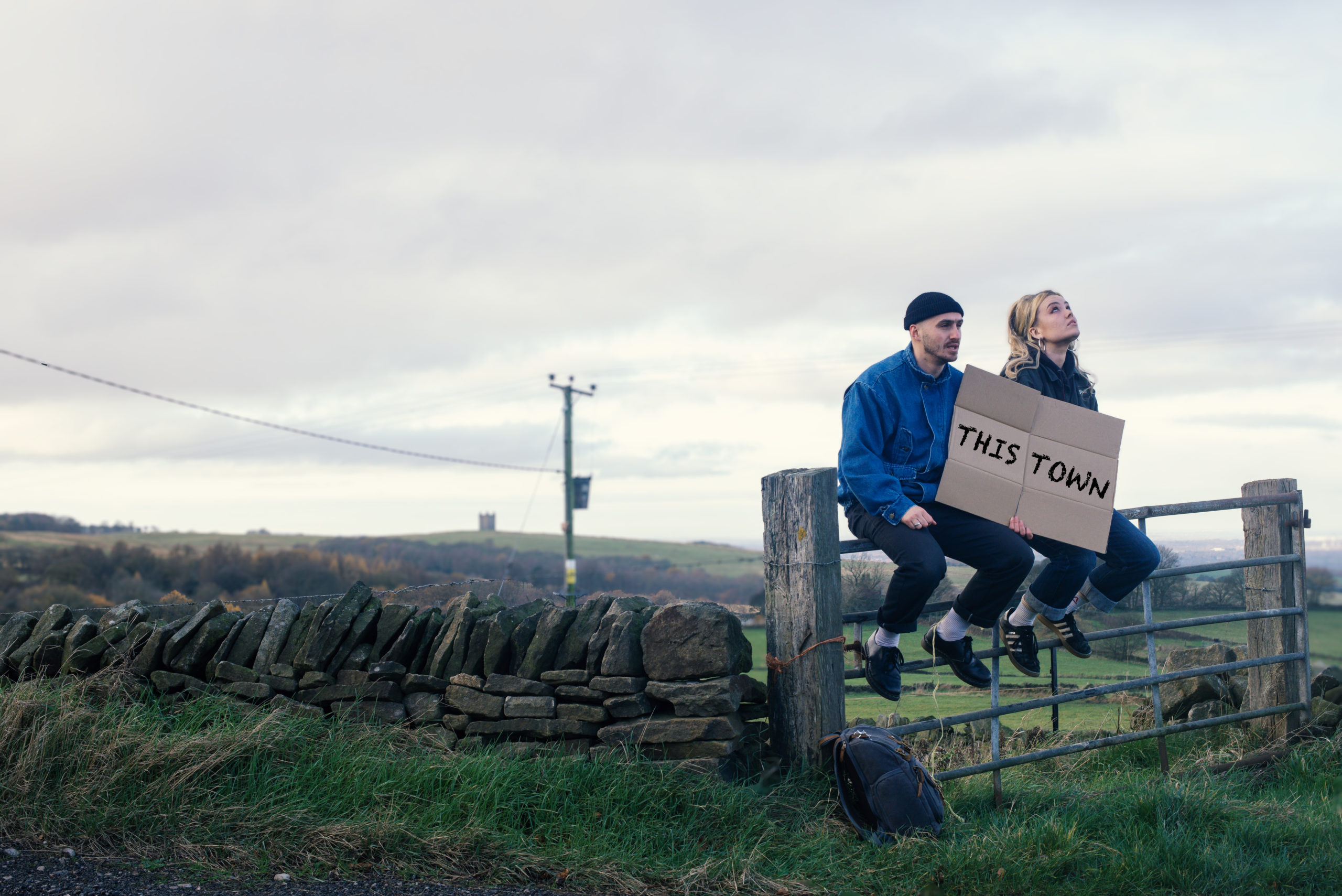 Two people sat on a fence with a cardboard sign that reads This Town